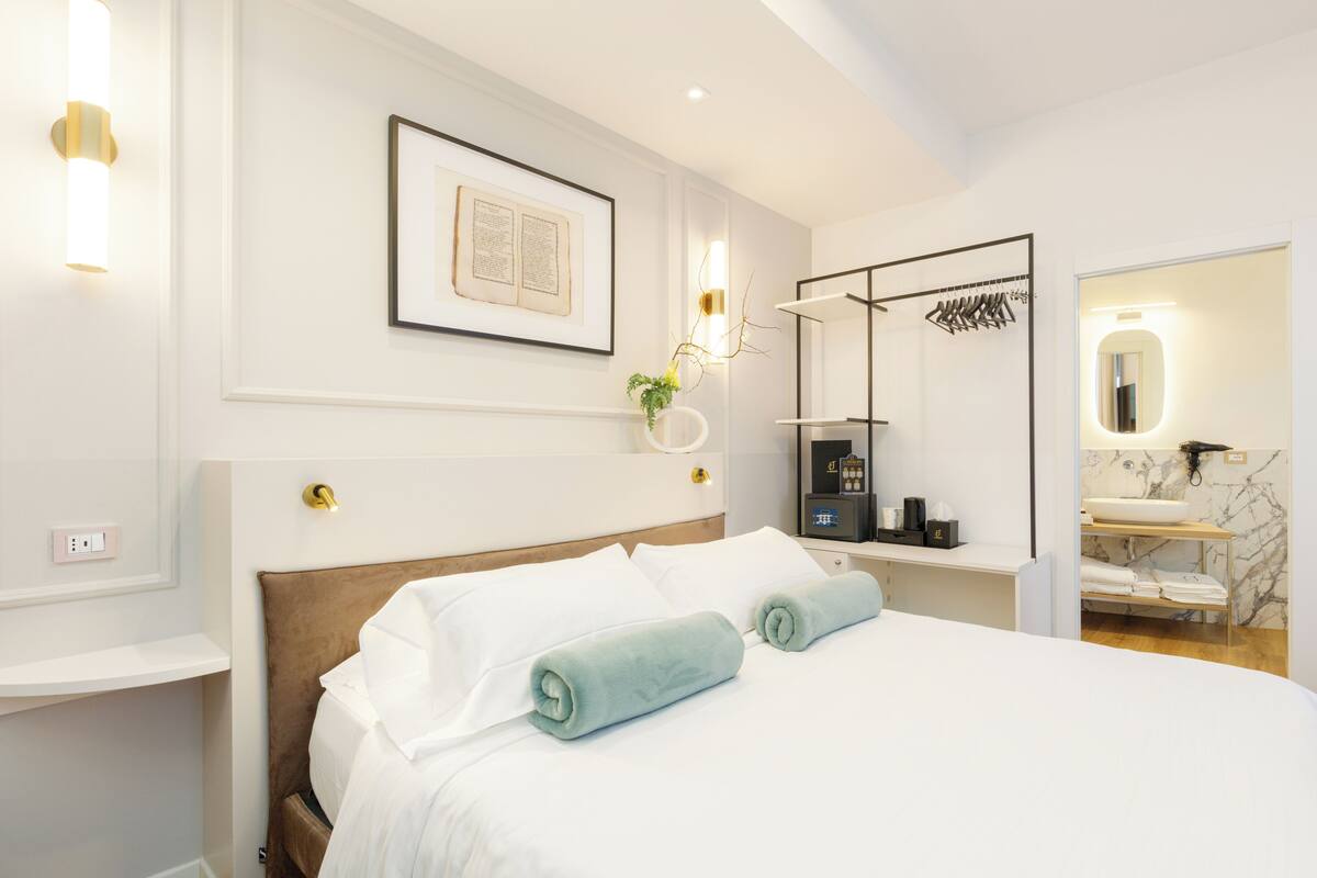 Une chambre d’hôtel comprenant un lit, un tableau encadré, une lampe fixée au mur, un miroir et une salle de bain avec un lavabo et un porte-serviettes.