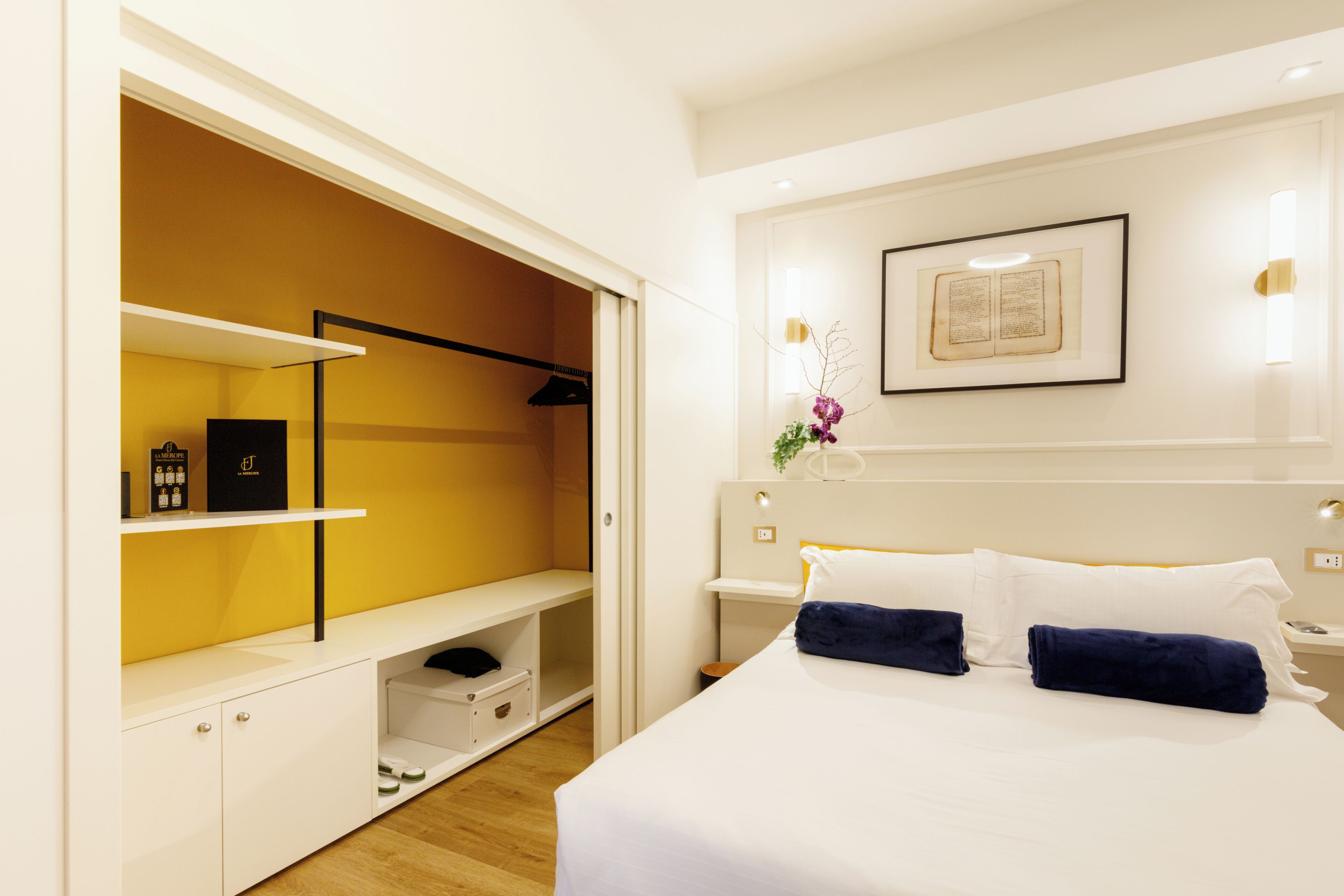 A hotel room with a bed, a nightstand, a bedside table, a wardrobe, and a framed picture on the wall.