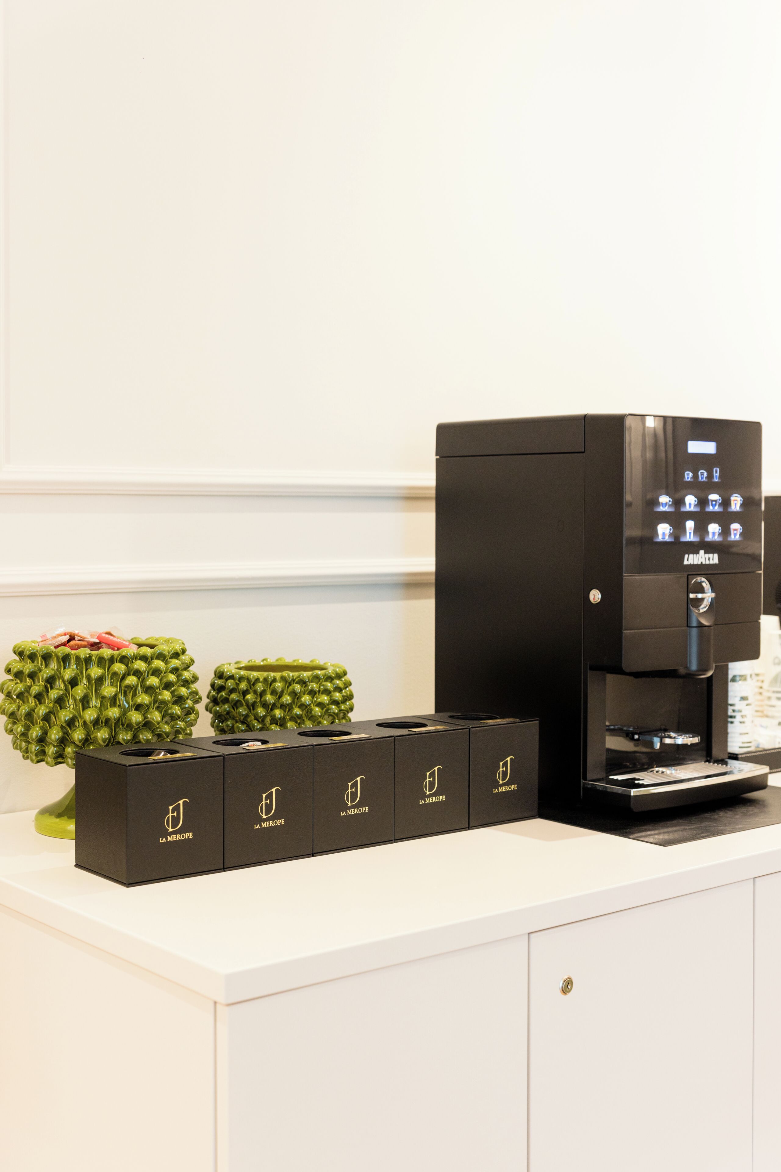 A coffee machine, four black boxes with a logo, and two green decorative items on a white counter.