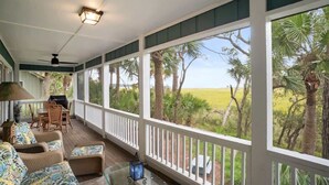 Eine überdachte Veranda mit Blick auf ein grasbewachsenes Feld und Palmen, möbliert mit Korbstühlen und einem Tisch.