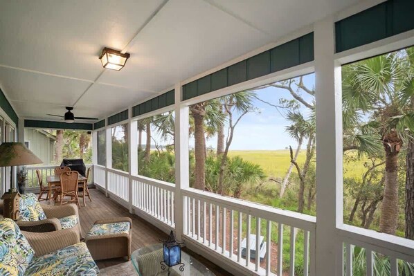 A covered porch with a view of a grassy field and palm trees, furnished with wicker chairs and a table. - 110 Sandpiper Run (Fripp Island)