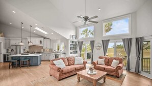 A spacious living area with a large window, a ceiling fan, and a kitchen in the background.