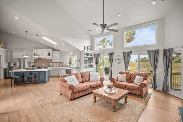 A spacious living area with a large window, a ceiling fan, and a kitchen in the background.