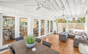 Un patio techado con mobiliario de mimbre, una mesa de madera con una planta y puertas correderas de cristal que dan a un espacio interior.