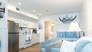 Una cucina moderna con mobili bianchi, un frigorifero in acciaio inossidabile e un forno a microonde. Nell'area notte, un letto con testiera blu decorata.