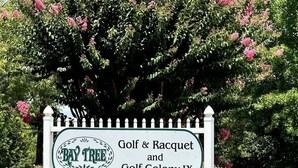 Schild für 'Bay Tree Golf & Racquet und Golf Colony IX' mit weißem Zaun und einem großen blühenden Baum im Hintergrund.