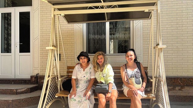 Three people sitting on a wooden swing bench with a metal frame, in front of a building with a brick facade and a window with decorative glass. - Sweet Home (Samarkand)
