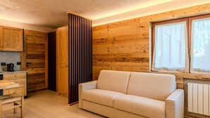 Un salon chaleureux avec un canapé beige, des murs en bois, une fenêtre avec des rideaux et un coin cuisine doté de meubles en bois et d’une machine à café.