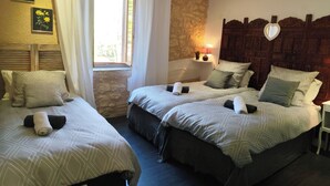 Ein Hotelzimmer mit zwei Betten, einer Steinwand, einem Holzkopfende, einem Fenster mit Vorhängen und einer Nachttischlampe.