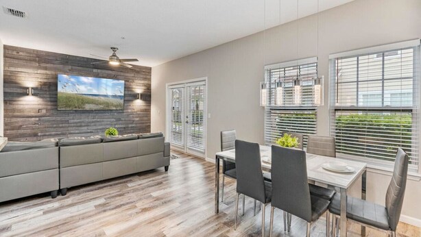 Un salon moderne avec un canapé gris, une table à manger avec des chaises, un téléviseur fixé à un mur en bois et de grandes fenêtres équipées de stores.