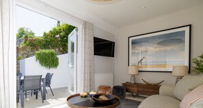 A modern living room with a dining area, a large window with a view of greenery, a flat-screen TV mounted on the wall, and a framed artwork depicting a beach scene. - Mudlark | Boutique Style Coastal Cottage Gem (Salcombe)