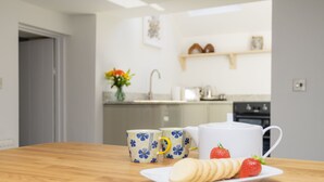 Una cocina con una mesa de madera, dos tazas con motivos florales, una tetera blanca y un plato con galletas y una fresa.