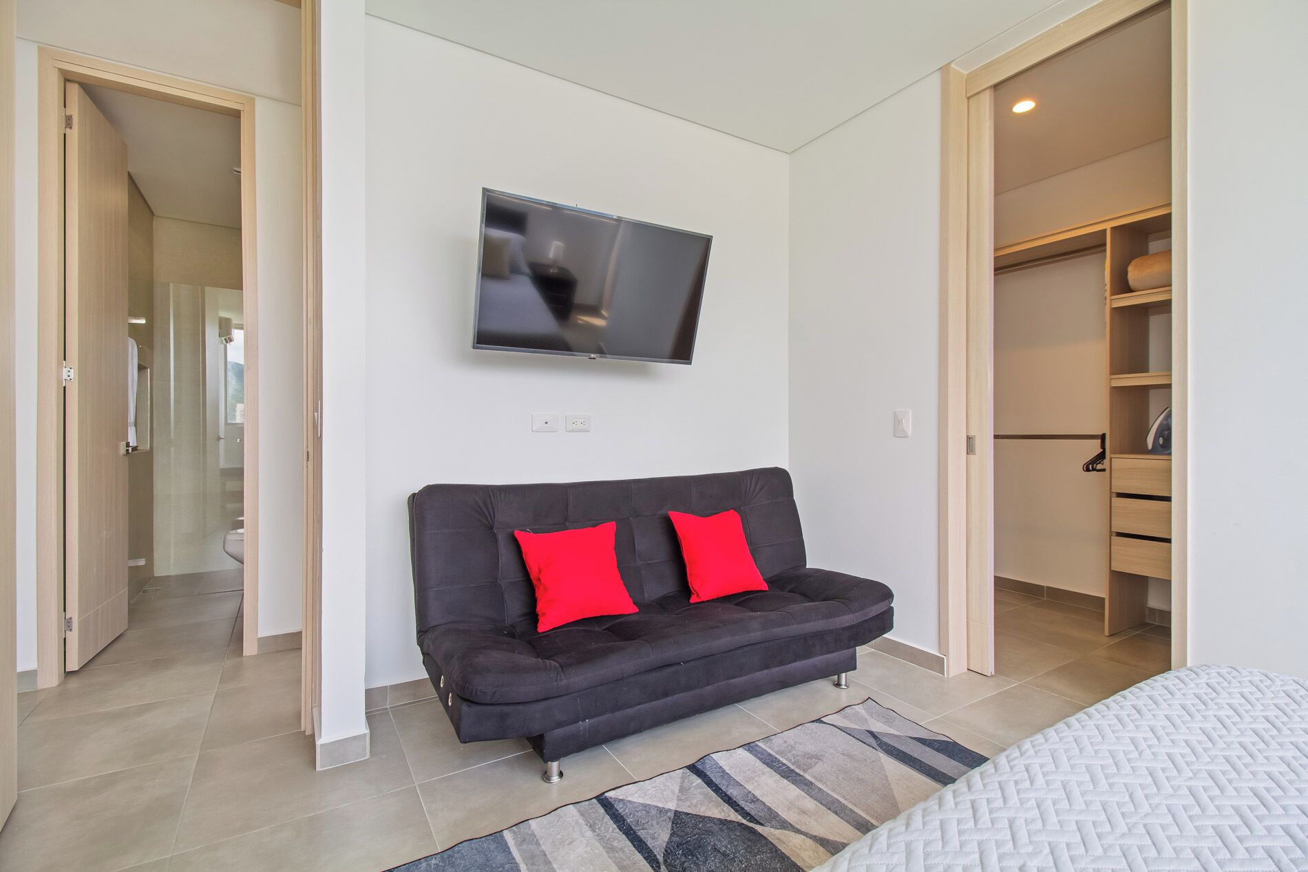 Une chambre moderne avec un canapé gris foncé, deux coussins rouges, un téléviseur à écran plat fixé au mur et un dressing avec des portes en bois.
