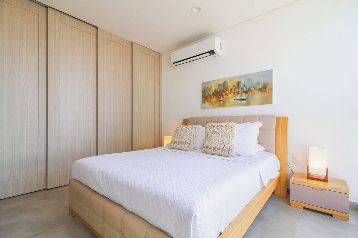 A bedroom with a bed, a nightstand, a painting, and an air conditioning unit.