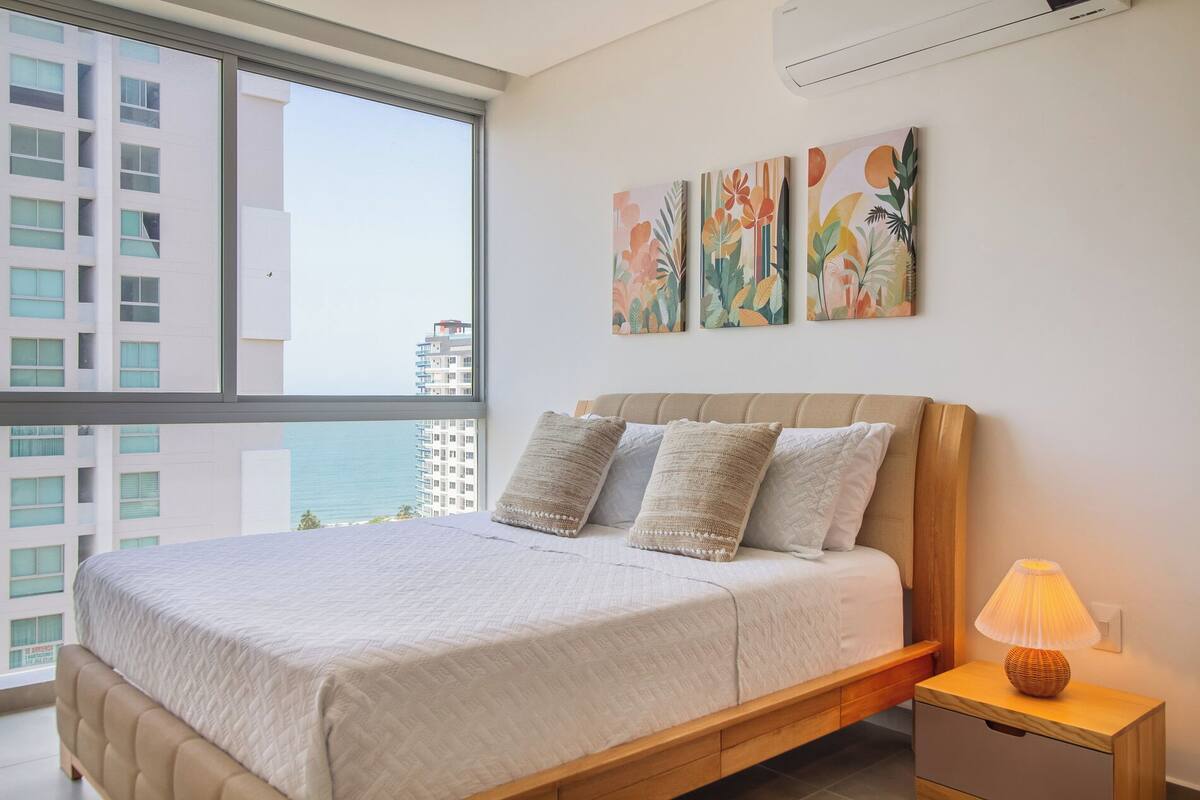 A bedroom with a bed, pillows, a nightstand, a lamp, and artwork on the wall.