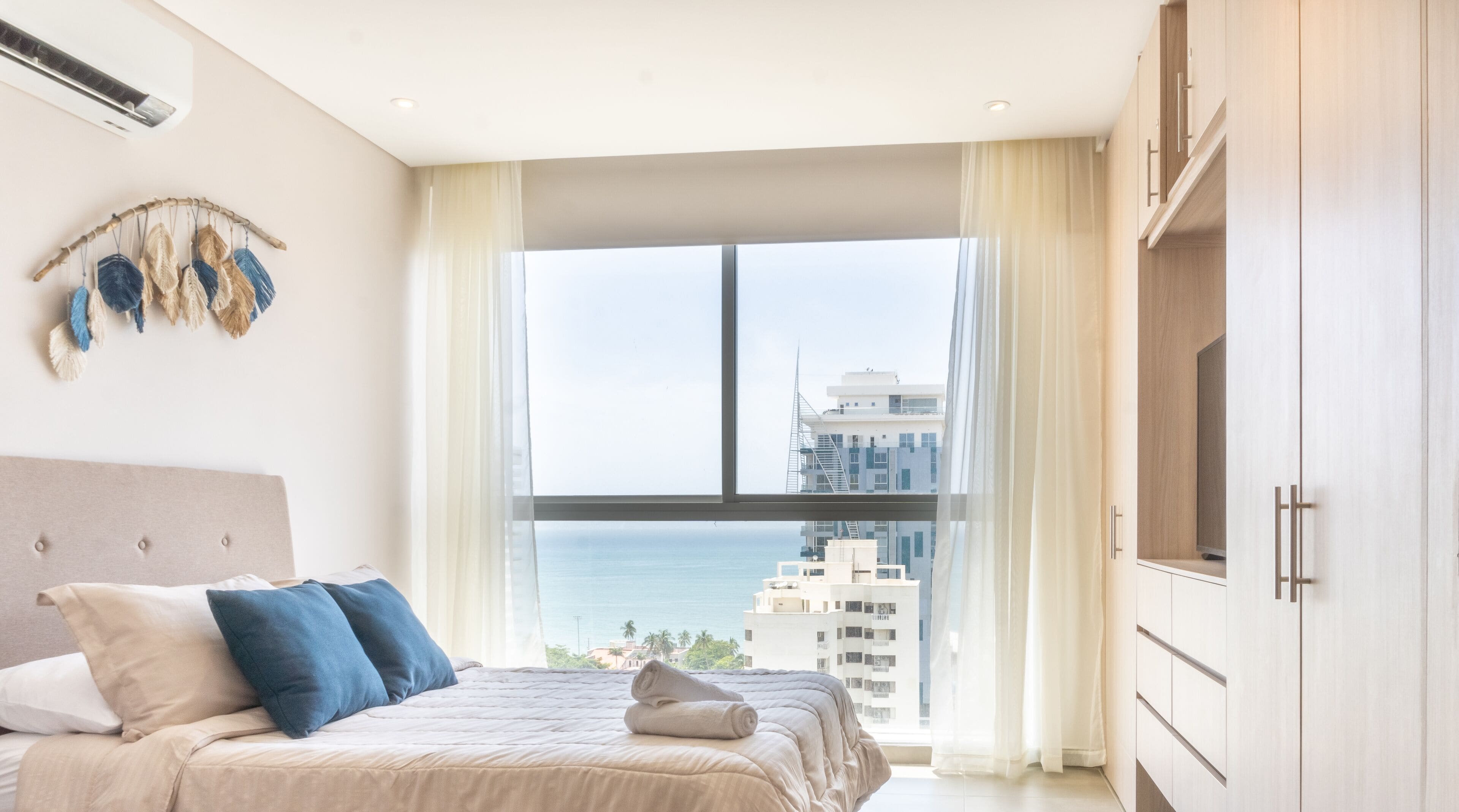 Un dormitorio moderno con un ventanal grande, una cama con ropa de cama azul y beige, un aire acondicionado instalado en la pared y vistas a la ciudad.