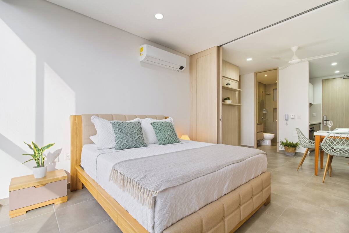 A modern bedroom with a large bed, a bedside table, a wardrobe, and an open-plan bathroom visible through a sliding door.