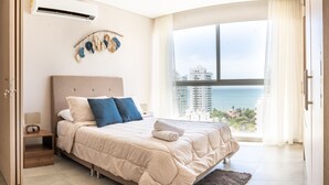 A bedroom with a large bed, a nightstand, an air conditioner, and a view of the ocean.