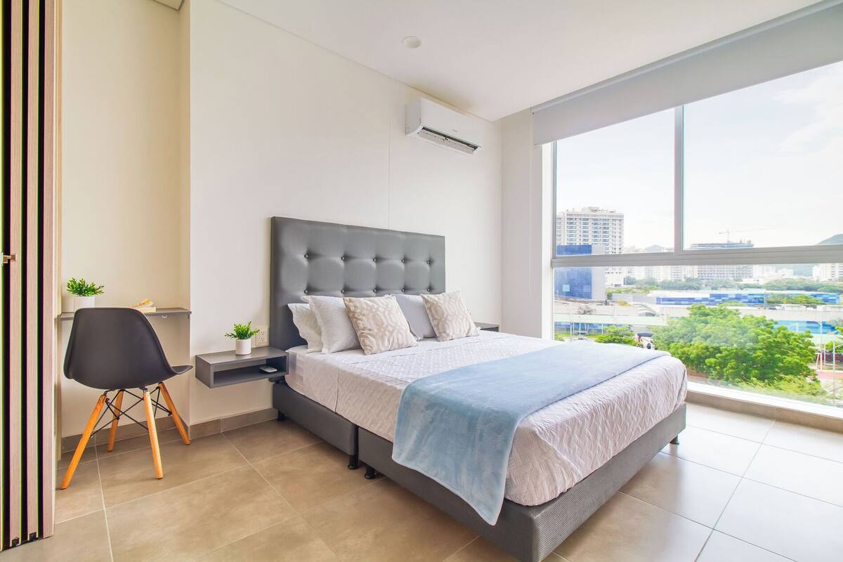 A modern bedroom with a large bed, a desk, and a chair. There is a window with a city view.