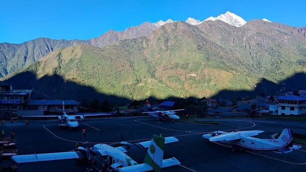 Un aeropuerto pequeño con varios aviones estacionados en la pista, rodeado de montañas y un cielo azul despejado.