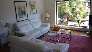 A living room with a white sofa, a glass coffee table, and a large window with a view of palm trees. - FERIENHAUS GERDA - Vacation in the sunniest region of Germany (Friesenheim)