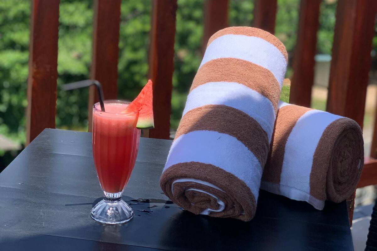 A wooden table with a glass of red drink and a rolled towel on a balcony overlooking a tropical landscape.