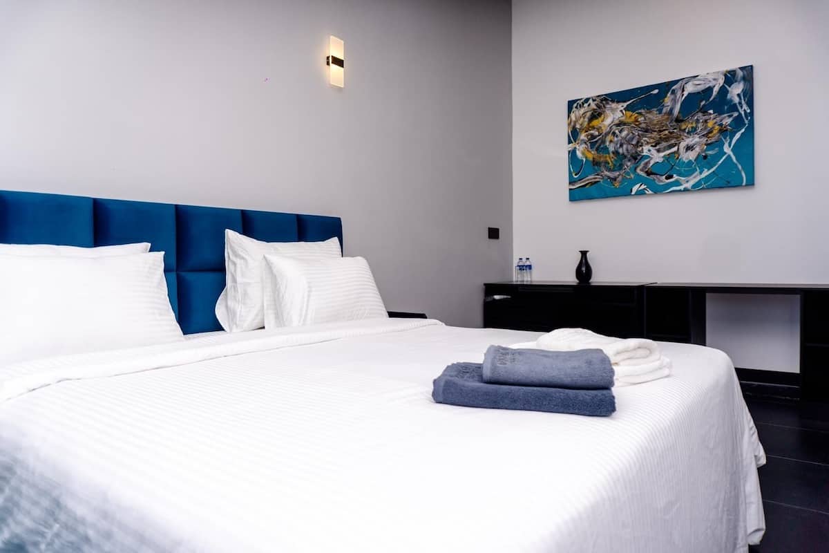 A modern hotel room with a large bed, a blue headboard, white bedding, a painting on the wall, and a black nightstand with a vase and water bottles.