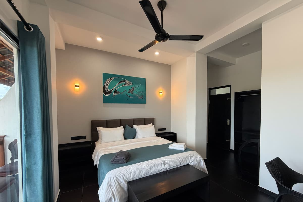 A modern hotel room with a large bed, a ceiling fan, a painting on the wall, and a view of the outside.