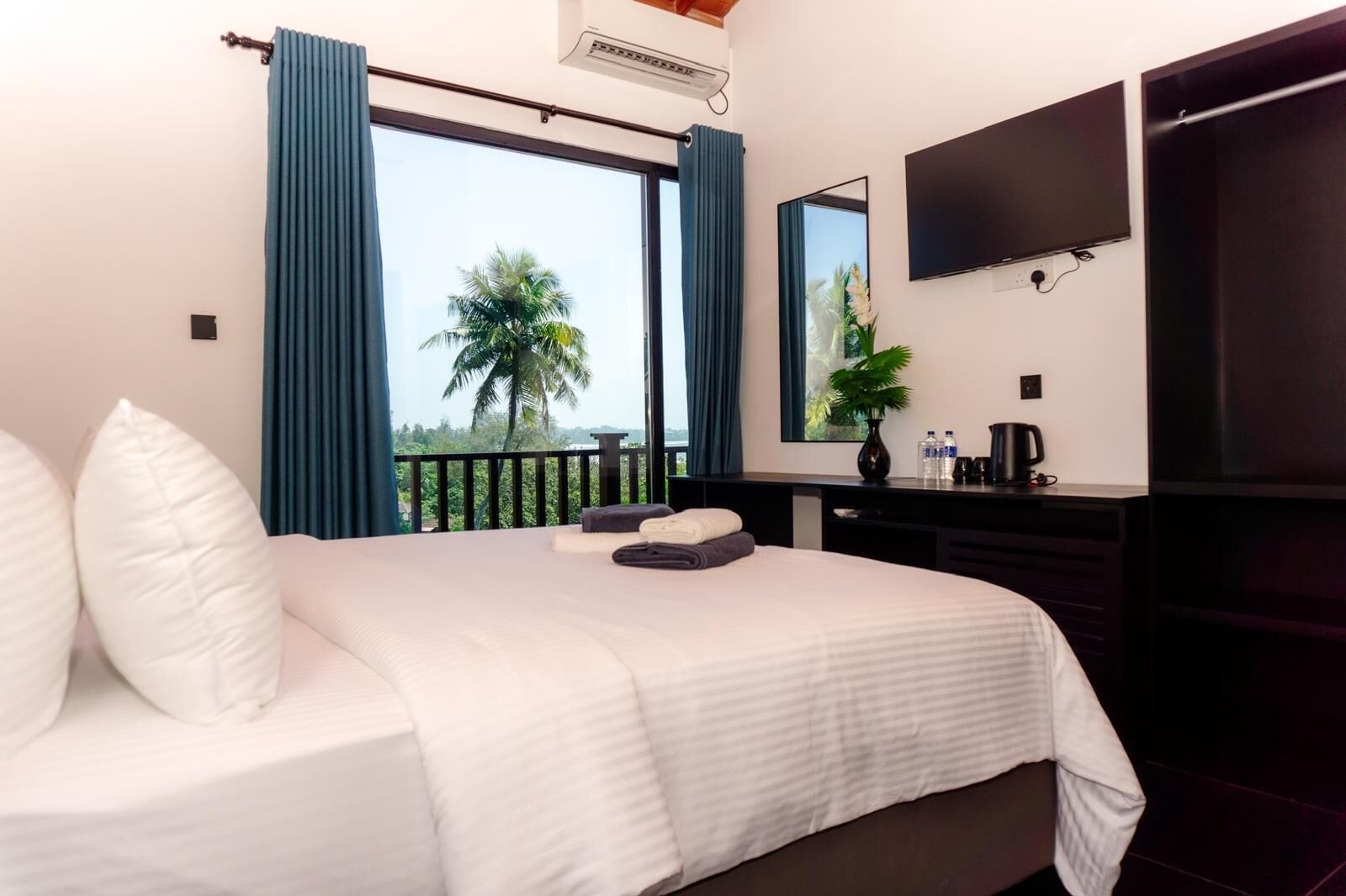 A hotel room with a large bed, a flat-screen TV, a desk with a coffee maker, and a view of palm trees through the window.