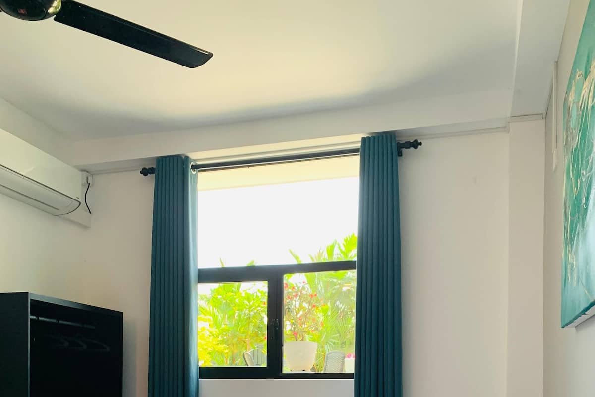A hotel room with two beds, a ceiling fan, a window with curtains, and a view of greenery outside.