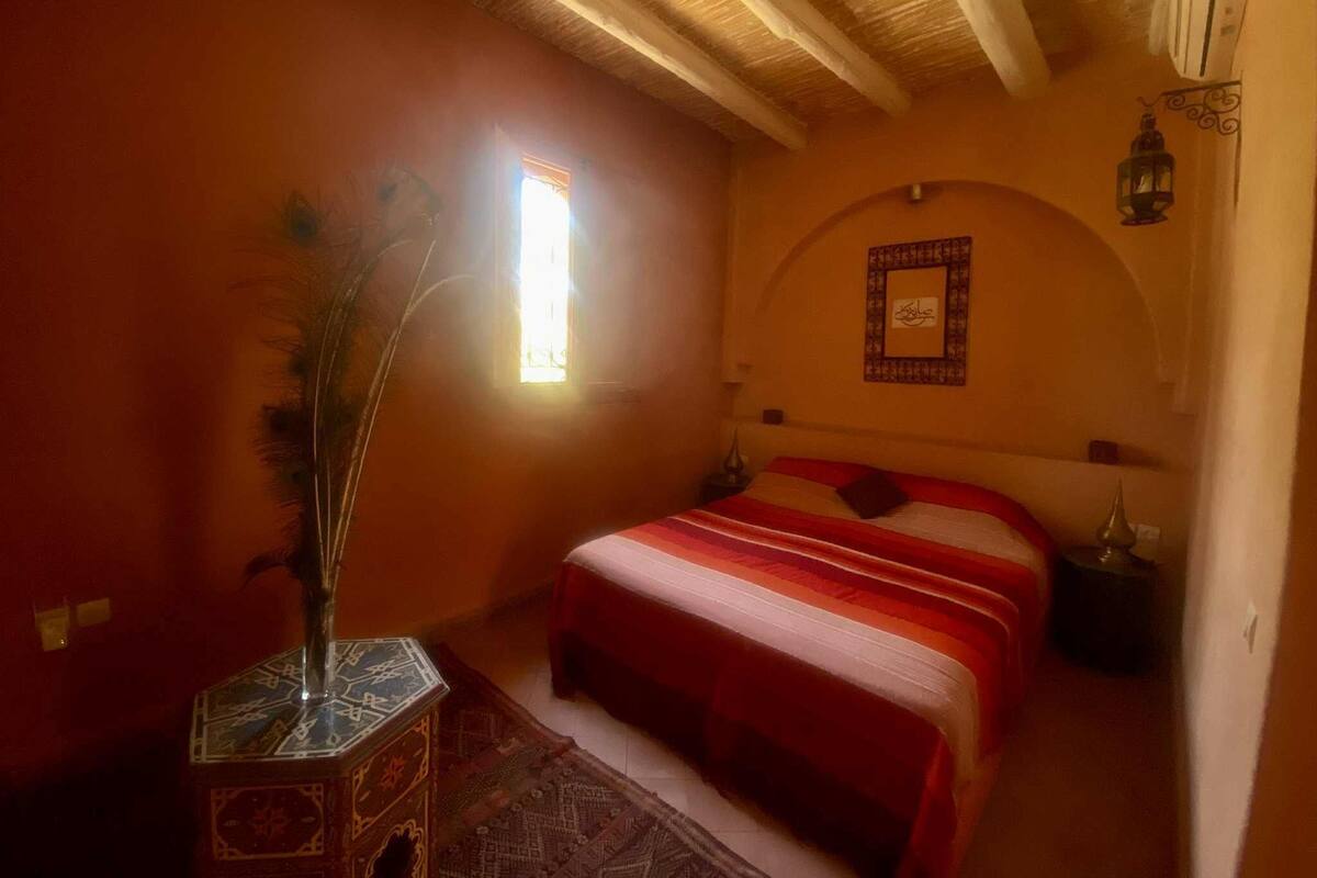 A bedroom with a bed, a window, a decorative wall piece, and a nightstand.