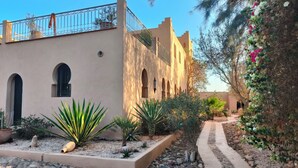 A courtyard with a pathway, potted plants, and a building with arches and a balcony. - Riad Jardin des Orangers (Sidi Borja)