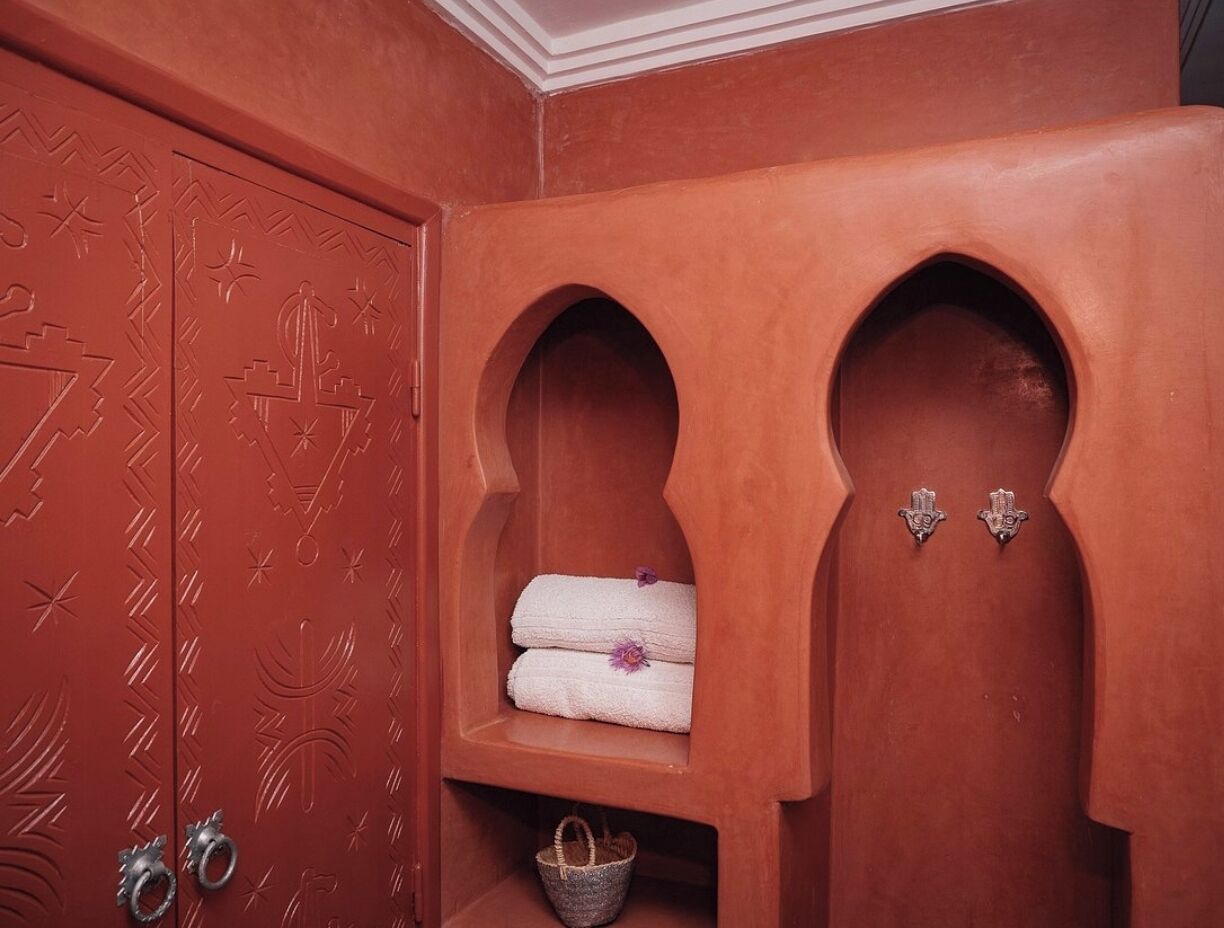 A red-tiled bathroom with a door featuring geometric patterns, a shelf with folded towels, and a small basket.