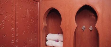 A red-tiled bathroom with a door featuring geometric patterns, a shelf with folded towels, and a small basket.