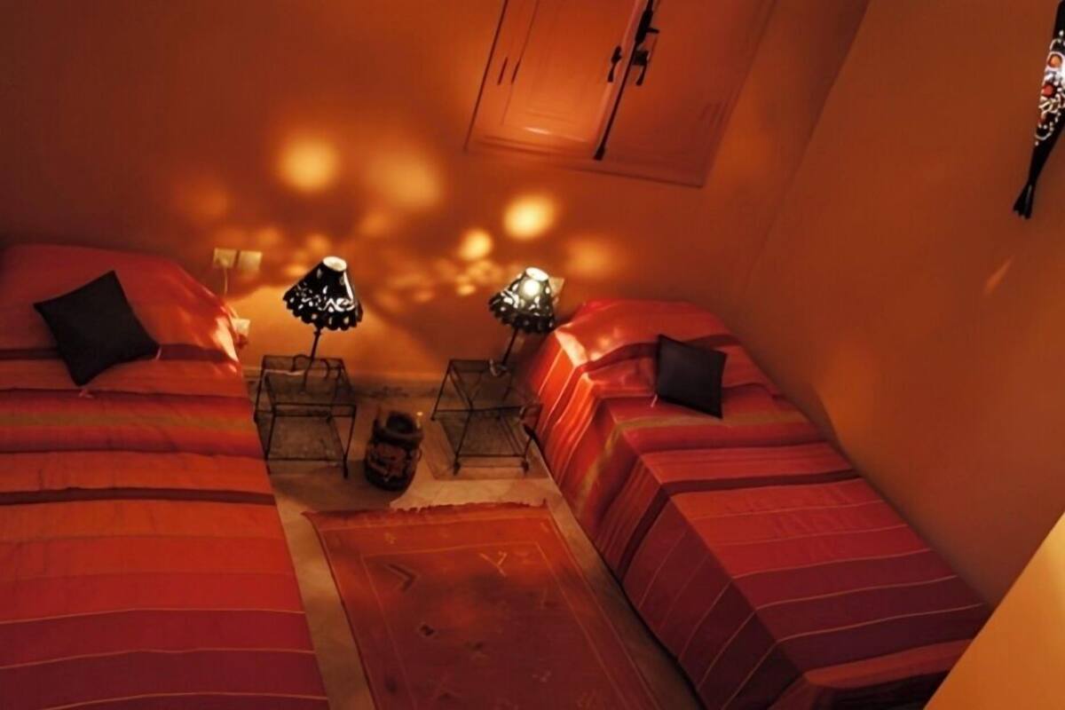 A cozy room with two beds, each with a red blanket and black pillows, a small table with a lamp, and a wall-mounted cabinet.