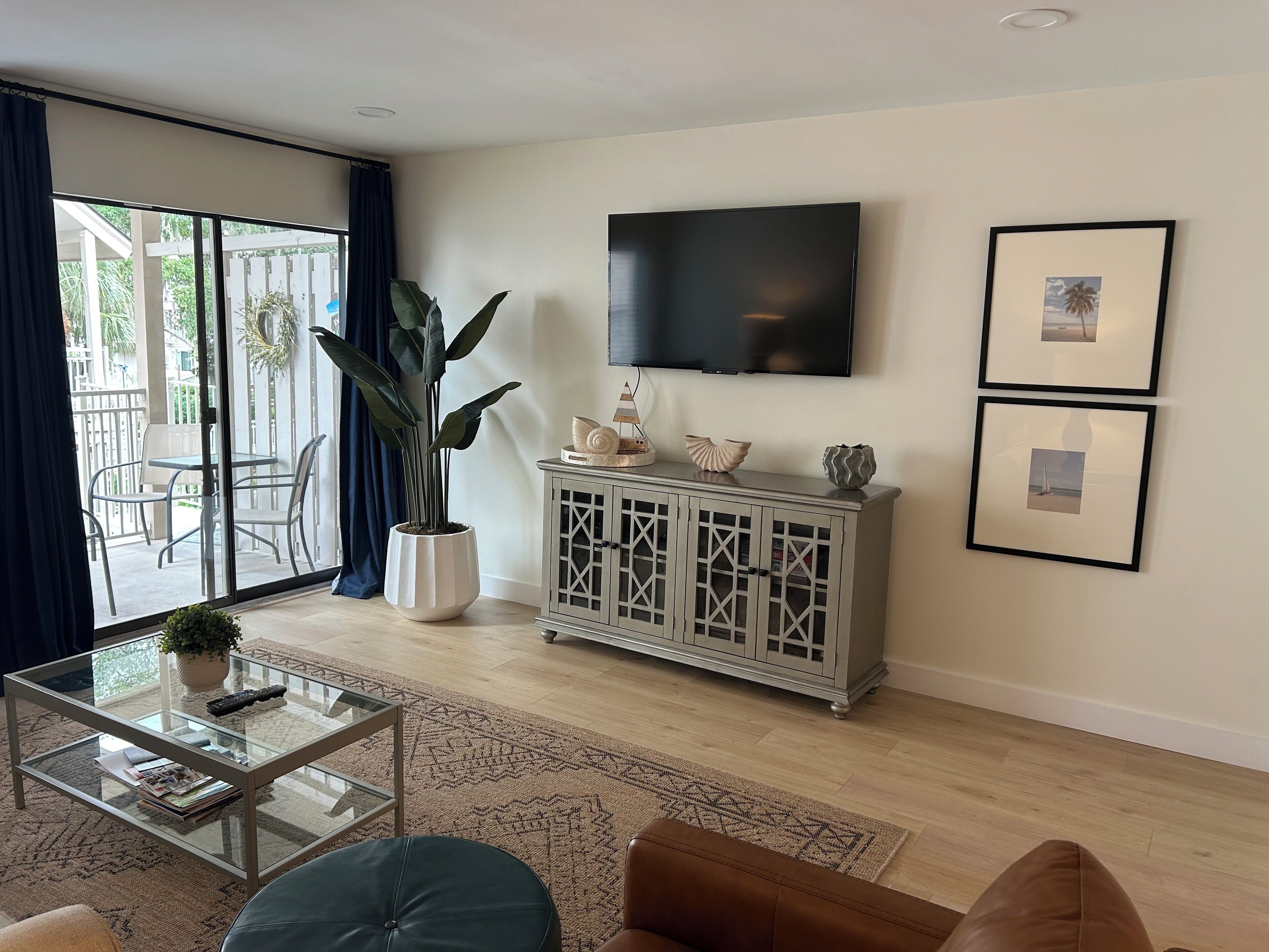 Ein Wohnzimmer mit Glastisch, Ledersofa, einem Topf mit Pflanze, einer Vitrine mit Dekoration und einem an der Wand befestigten Fernseher.