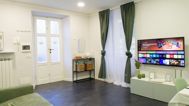 Een moderne woonkamer met een flatscreen-televisie die verschillende zenders toont. Er is een groene bank, een klein tafeltje met decoratieve items, en een donker houten vloer.