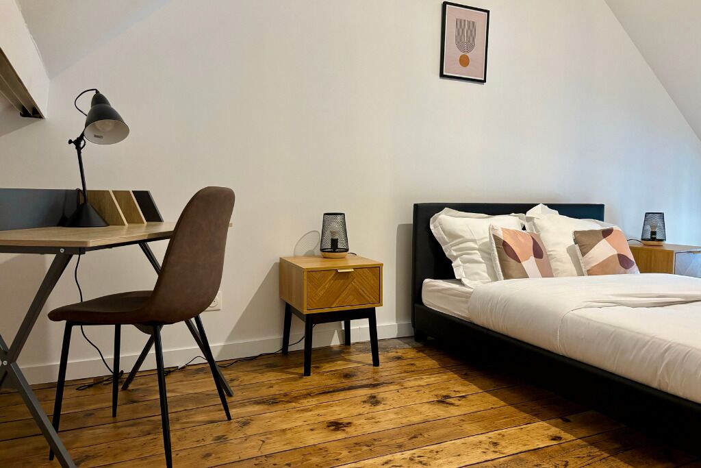 Een slaapkamer met een bed, een bureau met stoel, een kleine tafel met lamp, en een ingelijst kunstwerk aan de muur.
