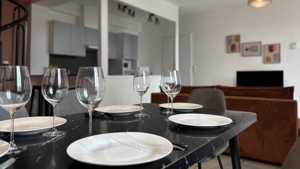 Une table à manger dressée avec des assiettes et des verres à vin, un canapé marron et, au fond, un coin cuisine.