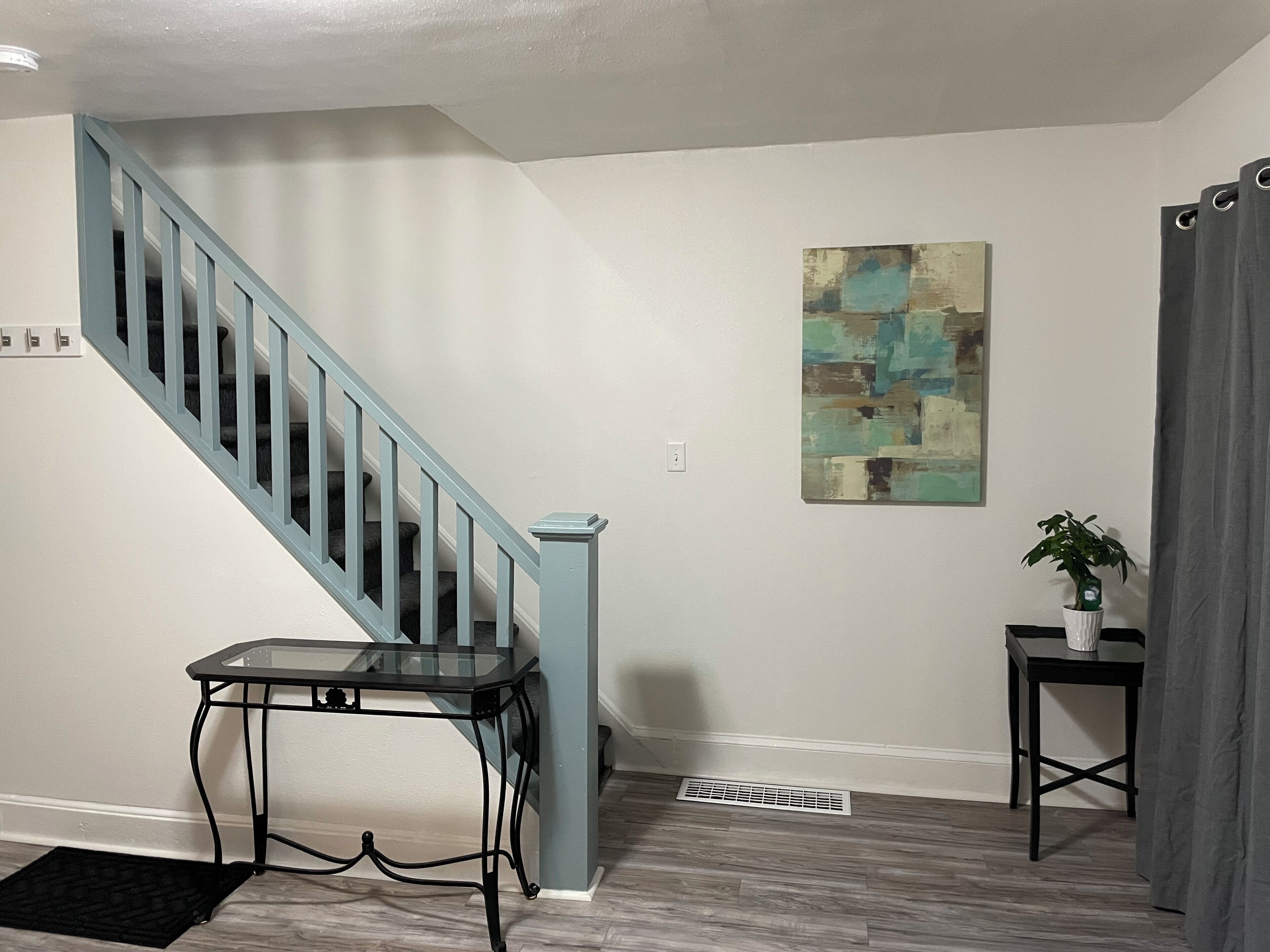 Un escalier avec des rampes bleu clair, une petite table avec un plateau en verre, un tableau accroché au mur, une plante en pot et un rideau gris.