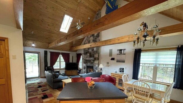 A spacious living area with a wooden ceiling, a stone fireplace, a dining table with chairs, and a kitchen island.
