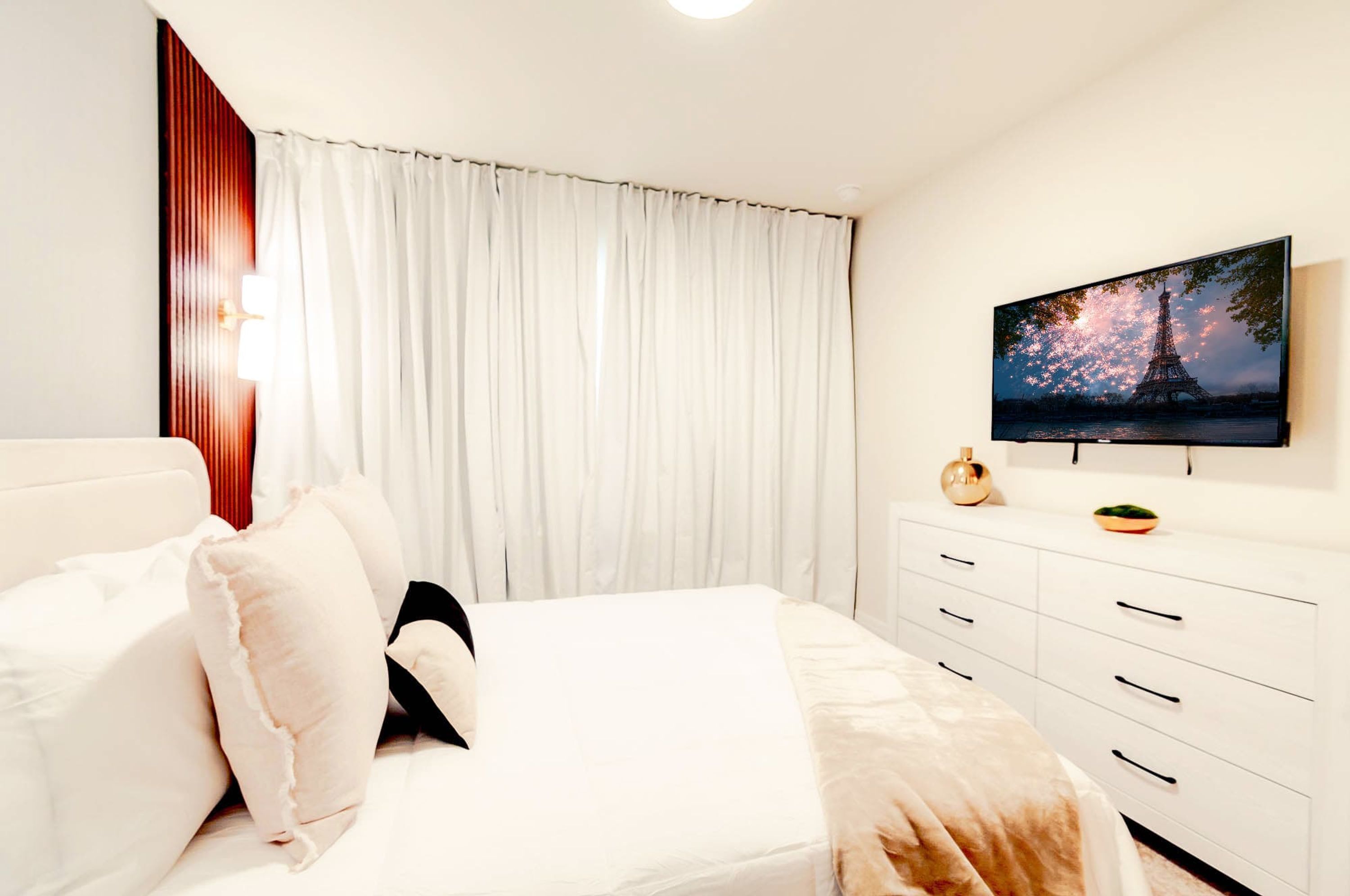 Une chambre d’hôtel moderne avec un grand lit, des rideaux blancs, un téléviseur à écran plat fixé au mur et un panneau en bois sur le côté gauche.
