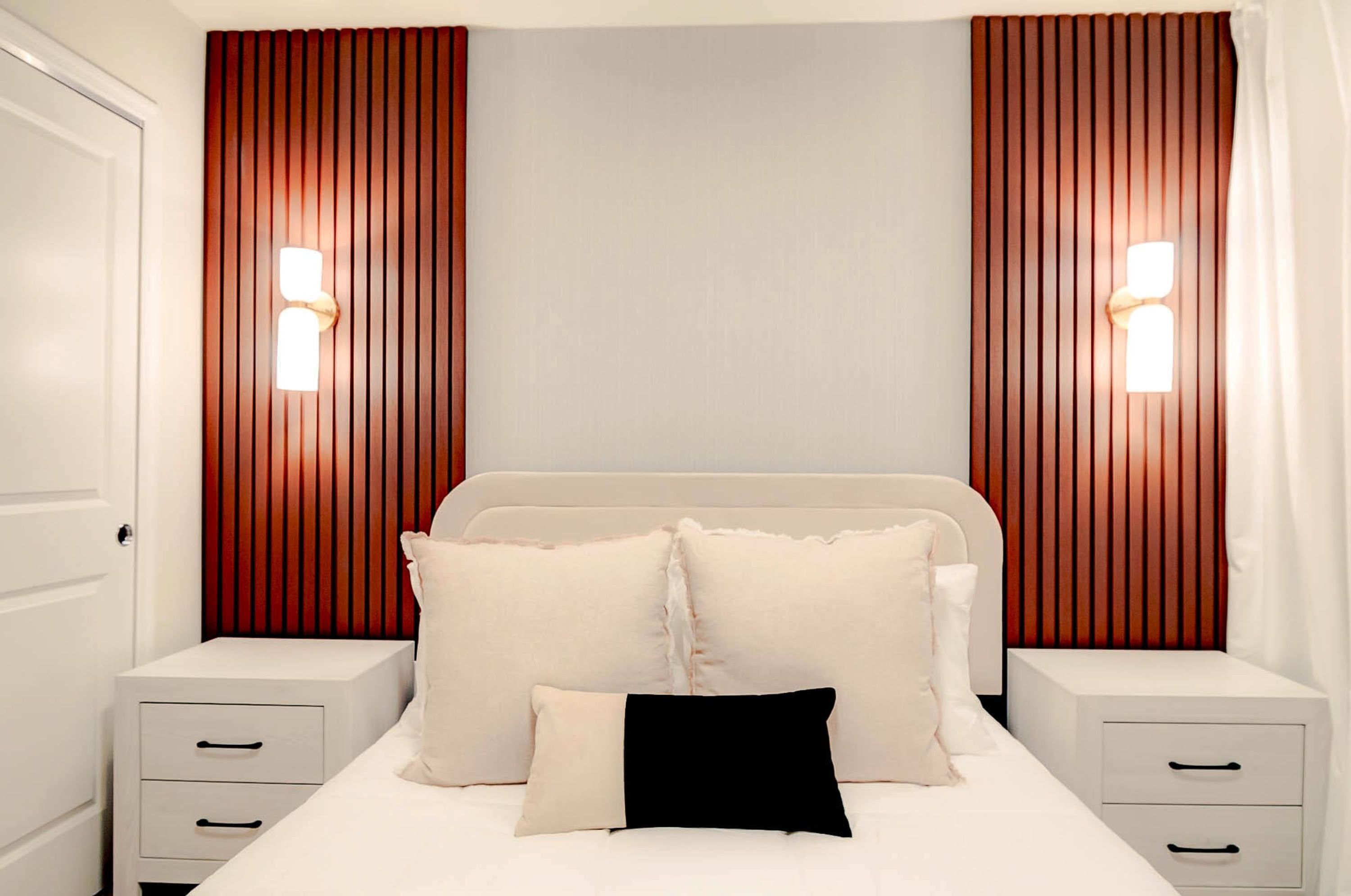 A bedroom with a bed, two bedside tables, a nightstand, and vertical wooden paneling with wall sconces.