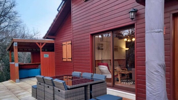 Un edificio de madera roja con un porche cubierto, una puerta de cristal que da a un espacio interior y mobiliario exterior.