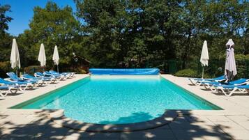 A rectangular swimming pool with clear blue water, surrounded by white lounge chairs and umbrellas, set against a backdrop of trees.