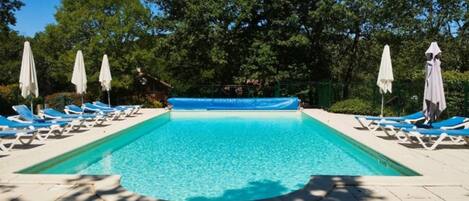 A rectangular swimming pool with clear blue water, surrounded by white lounge chairs and umbrellas, set against a backdrop of trees.