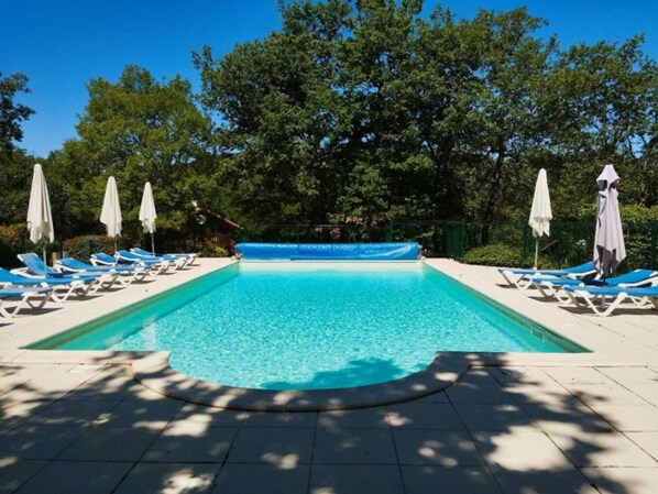 A rectangular swimming pool with clear blue water, surrounded by white lounge chairs and umbrellas, set against a backdrop of trees.