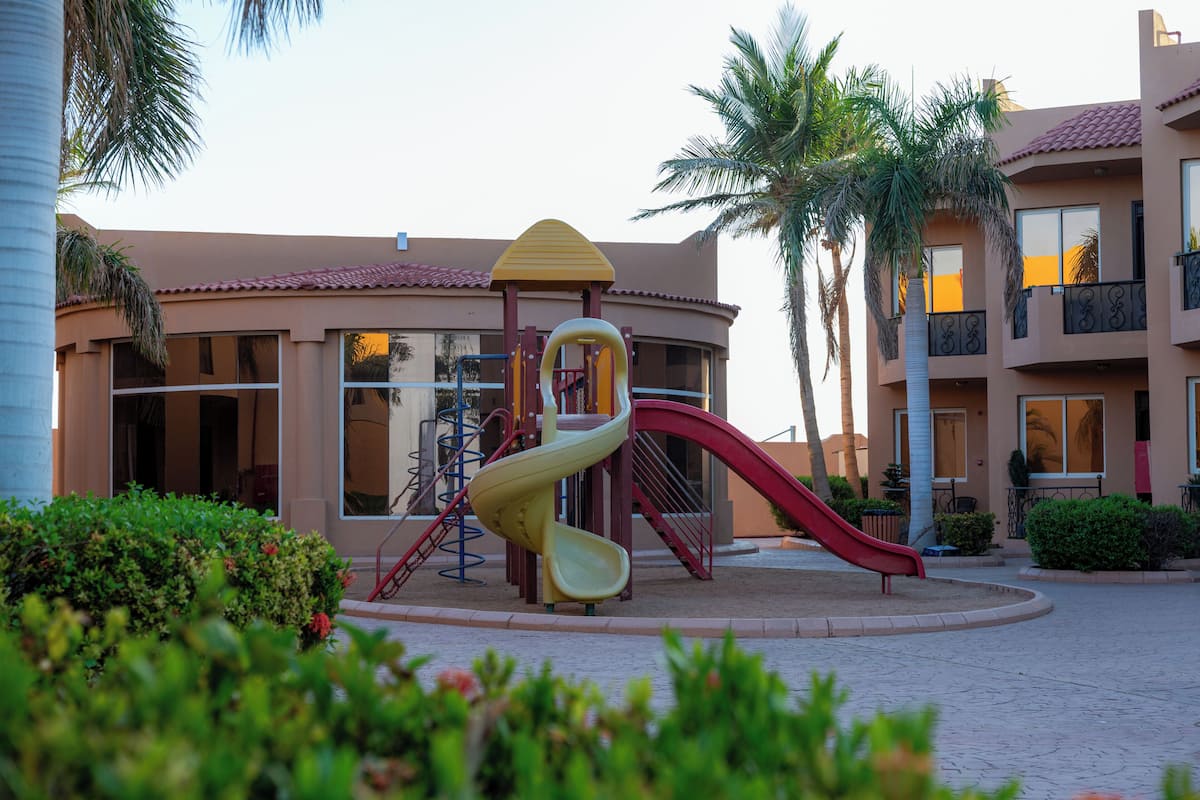 A playground with slides and climbing structures, surrounded by palm trees and a building with balconies.