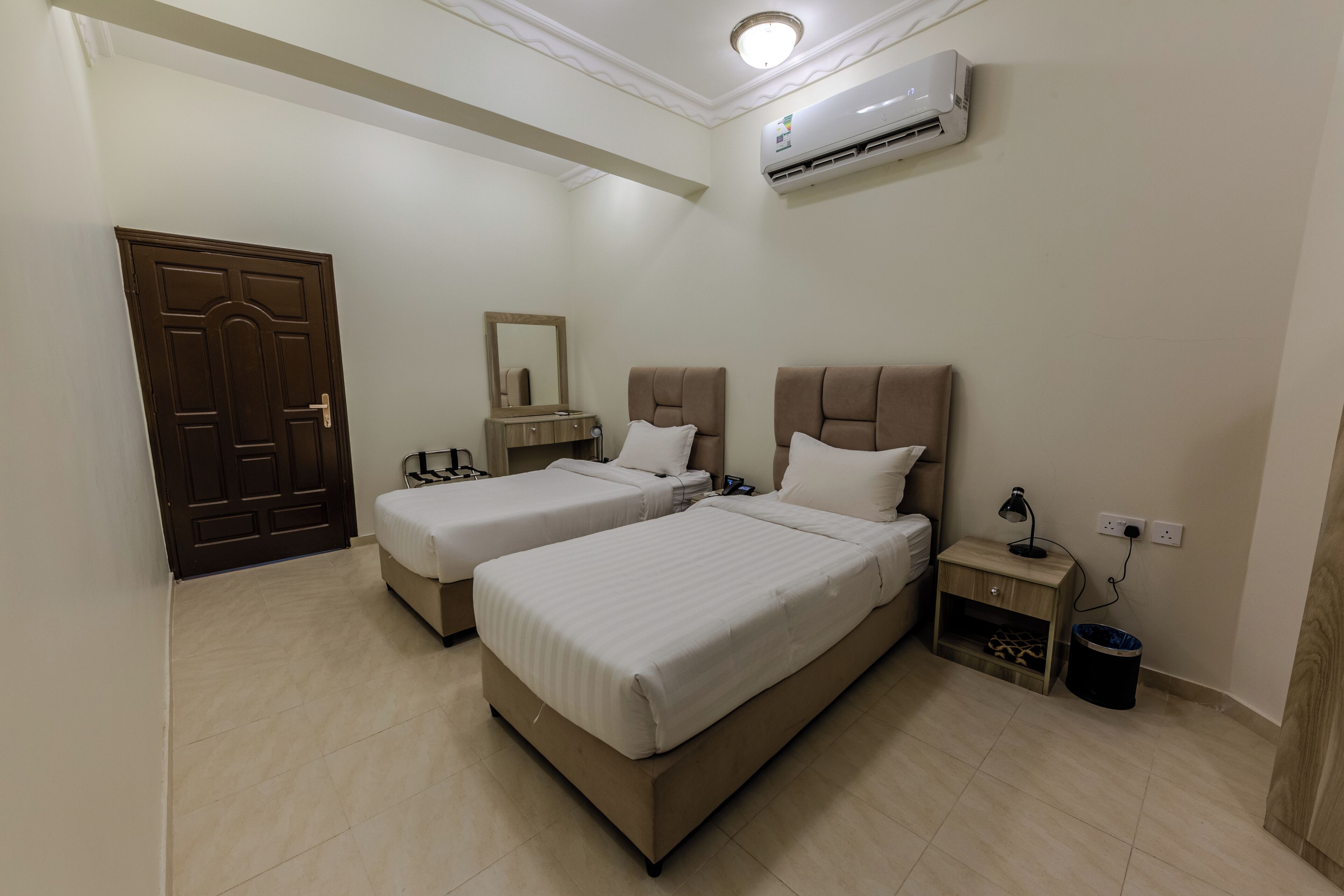 A hotel room with two beds, a wardrobe, a mirror, an air conditioner, a nightstand with a lamp, and a trash bin.