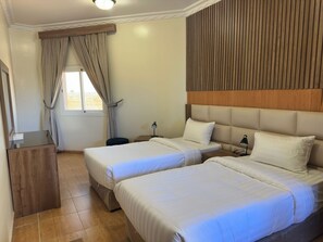 Two single beds with white linens and a wooden headboard, a desk, and a window with curtains.